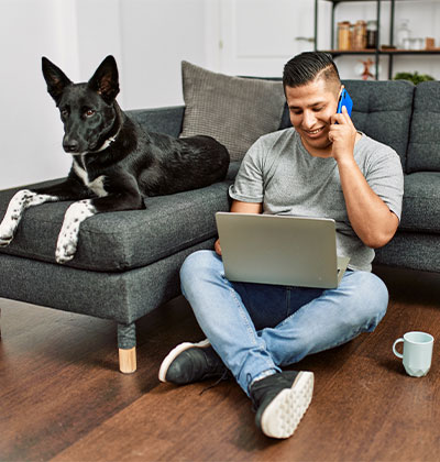 A man sits on the floor, leaned against a dark grey couch, talking to his veterinarian on the phone while using a laptop. A black and white dog is lying on the couch behind him. The scene includes a coffee mug on the wooden floor.
