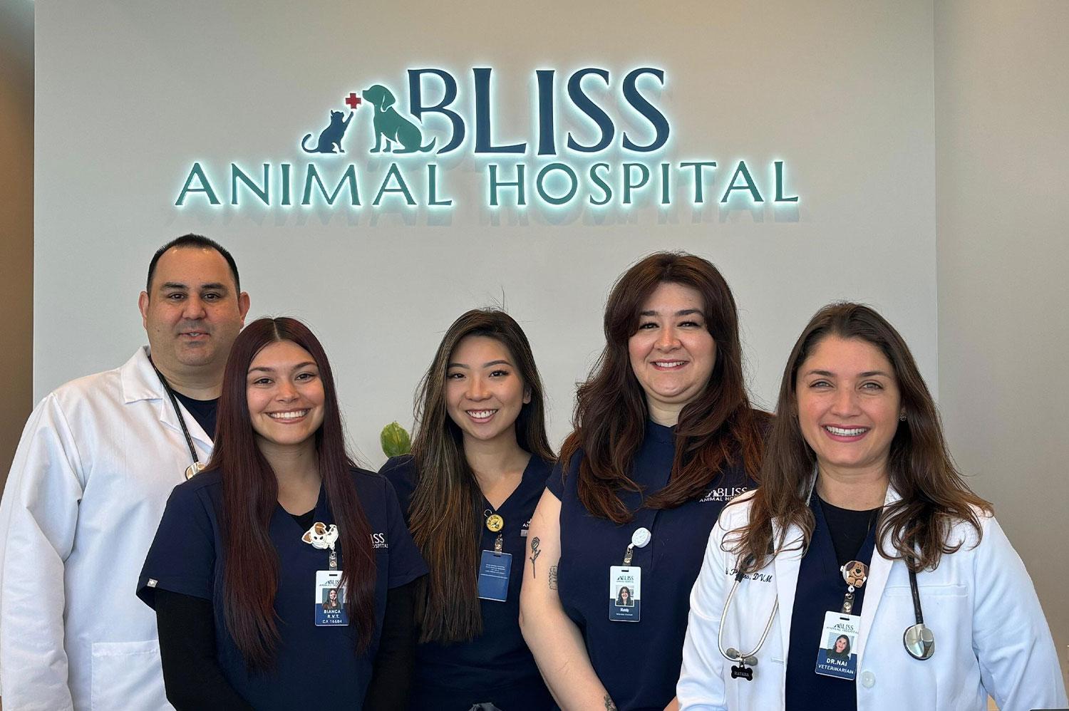 Five staff members of Bliss Animal Hospital are standing together and smiling at the camera. Three women in navy uniforms and one veterinarian in a doctor's coat stand in front of a sign that reads "Bliss Animal Hospital." Another male vet in a white uniform stands beside them.