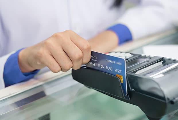 A person swipes a credit card through a point-of-sale terminal on a glass counter, likely at an affordable vet clinic, wearing a white long-sleeve shirt with blue cuffs.